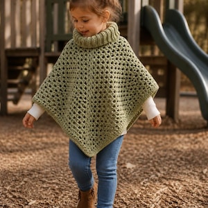 Könnte beinhalten: Ein salbeigrüner Häkelponcho mit einem dicken, gerippten Rollkragen und einem offenen Rautenmuster. Das Kleidungsstück wird über einem weißen Langarmshirt, einer blauen Jeanshose und braunen Wildleder-Stiefeletten mit Fransen getragen.