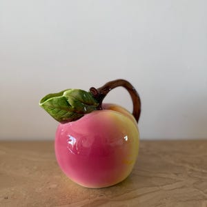 May include: A ceramic creamer shaped like a peach with a glossy finish, featuring a gradient of pink and yellow colours. The handle is designed to look like a brown tree branch, and a green leaf is attached near the spout of the pitcher.