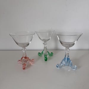 May include: Three clear glass martini glasses with colored stems. One stem is pink, one is green, and one is blue. Each glass has a triangular bowl and a three-footed base.