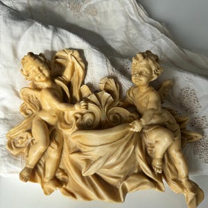 May include: A cream-coloured cherub sculpture featuring two winged figures holding a draped cloth. The cherubs have detailed facial features and are set against a backdrop of a white, textured fabric. The sculpture is a decorative piece.