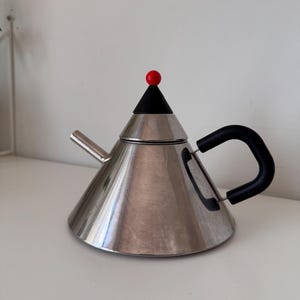 May include: A stainless steel teapot with a black handle and a conical lid topped with a red sphere. The teapot has a spout and sits on a white surface.