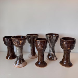 May include: Six handmade ceramic goblets with a dark brown glaze. The goblets have a pedestal base and a wide, open bowl. Some have a lighter, marbled effect on the stem. The goblets are arranged on a white surface.