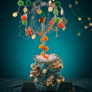 May include: A decorative wire tree sculpture with gold coins, keys, and pom-poms in a mosaic glass jar. The tree has a silver wire trunk and branches, with green, orange, and gold accents. The jar is teal with gold frog embellishments.