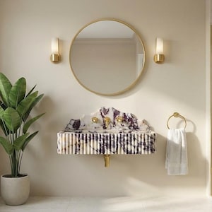 Puede incluir: Interior de baño con lavabo de mármol, grifería dorada y espejo redondo dorado. El lavabo tiene un patrón blanco y morado. Una planta en maceta está a la izquierda. Una toalla blanca cuelga de un toallero dorado.