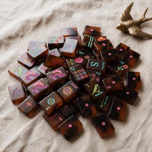 May include: A collection of rectangular mahjong tiles with a brown tortoiseshell pattern, featuring various engraved symbols, numbers, and floral designs in vibrant colors like pink, green, and blue, alongside a small brass bird figurine.