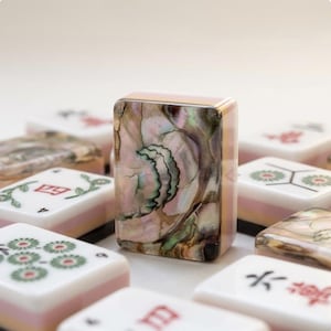 May include: A collection of rectangular mahjong tiles featuring a central tile with an iridescent, multi-colored abalone shell inlay. Surrounding tiles display white surfaces with red and green engraved symbols, numbers, and floral patterns.