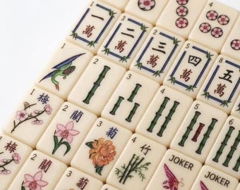 May include: A collection of off-white mahjong tiles arranged in a grid. The tiles feature various designs including Chinese characters, green bamboo stalks, colorful floral illustrations, and tiles labeled with the word JOKER in black text.