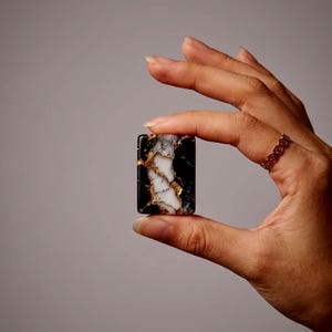 May include: A rectangular cabochon featuring a marbled pattern of black and white with metallic gold veining throughout the surface. The piece has a smooth, polished finish and a distinct contrast between the dark and light color sections.