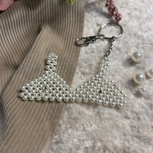 Puede incluir: Un llavero con forma de sujetador hecho de perlas blancas, con un cierre y una cadena plateados. El dise&ntilde;o del sujetador est&aacute; hecho de peque&ntilde;as perlas redondas. Fondo beige y blanco.