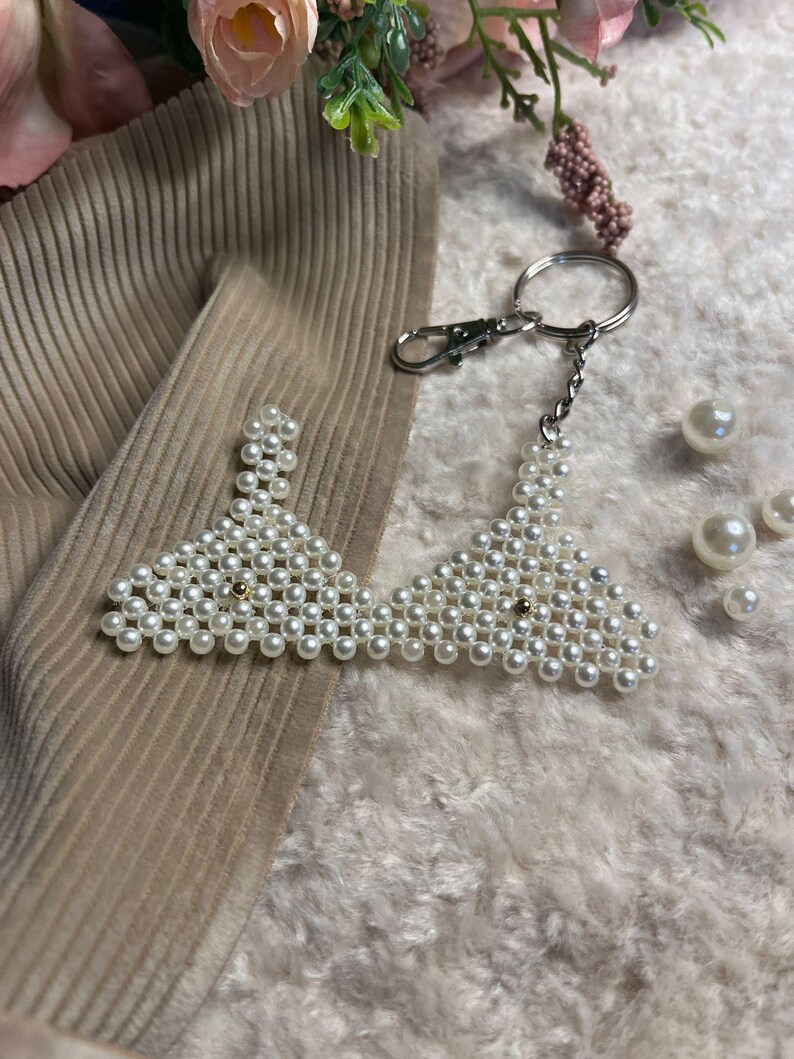 Puede incluir: Un llavero con forma de sujetador hecho de perlas blancas, con un cierre y una cadena plateados. El dise&ntilde;o del sujetador est&aacute; hecho de peque&ntilde;as perlas redondas. Fondo beige y blanco.