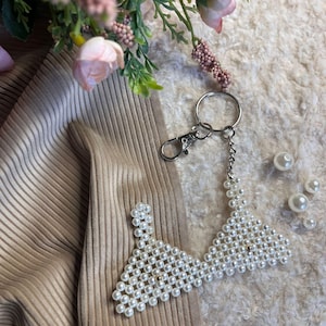 Può includere: Un portachiavi a forma di reggiseno con perline di perle. Il portachiavi ha un moschettone e una catena argentati. Il reggiseno è fatto di perle bianche e ha una perla dorata al centro. Sfondo strutturato con fiori rosa.