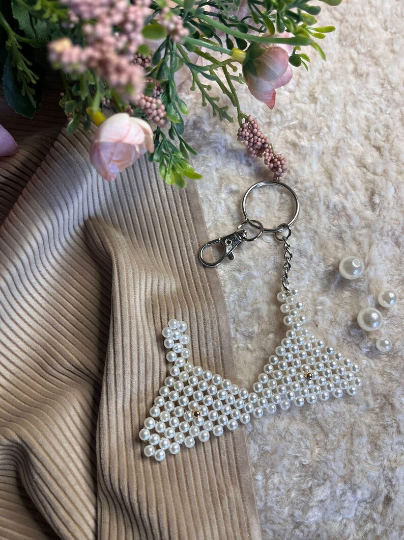 Puede incluir: Un llavero con cuentas de perlas en forma de sujetador. El llavero tiene un cierre y una cadena plateados. El sujetador est&aacute; hecho de perlas blancas y tiene una cuenta dorada en el centro. Fondo texturizado con flores rosas.