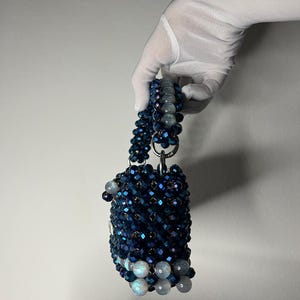 May include: A small, beaded handbag in dark blue and iridescent white. The bag has a cylindrical shape with a beaded handle. The bag is held by a gloved hand. The beads create a textured, sparkling effect, suitable for a special occasion.
