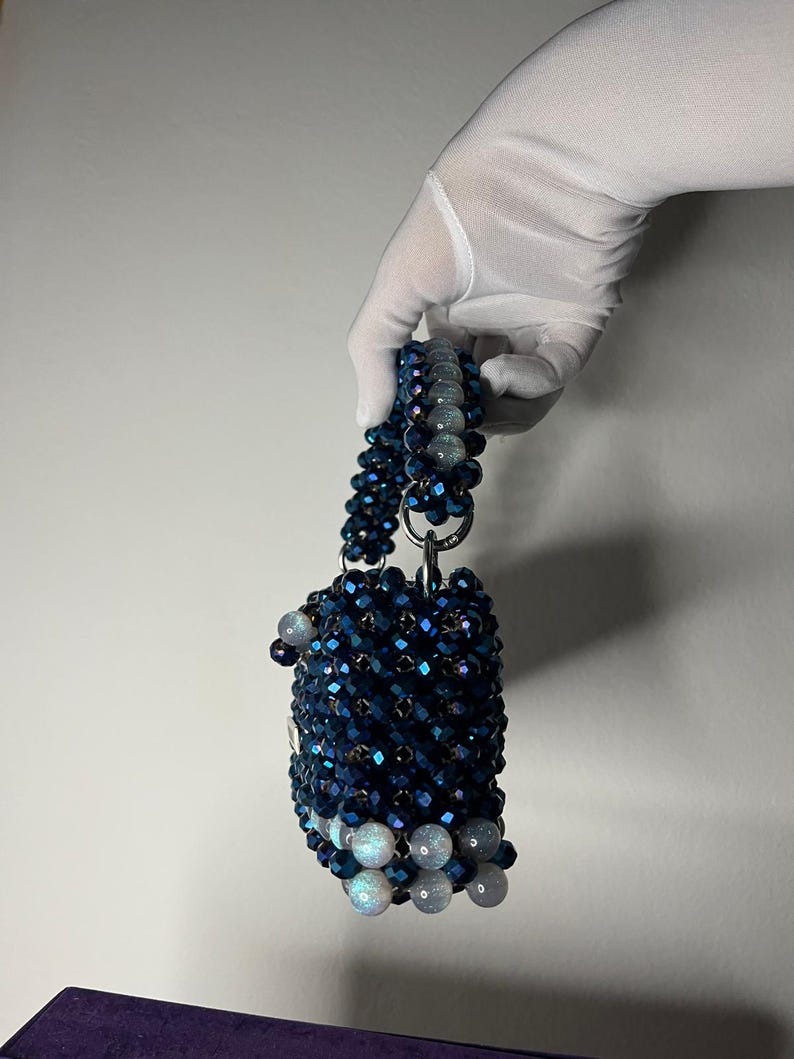 May include: A small, beaded handbag in dark blue and iridescent white. The bag has a cylindrical shape with a beaded handle. The bag is held by a gloved hand. The beads create a textured, sparkling effect, suitable for a special occasion.