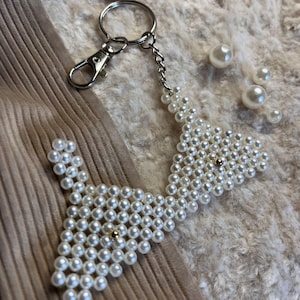 Puede incluir: Un llavero blanco con cuentas en forma de pajarita con un cierre y cadena plateados. El llavero est&aacute; hecho de peque&ntilde;as cuentas blancas redondas y presenta una peque&ntilde;a cuenta dorada en el centro de cada secci&oacute;n de pajarita.