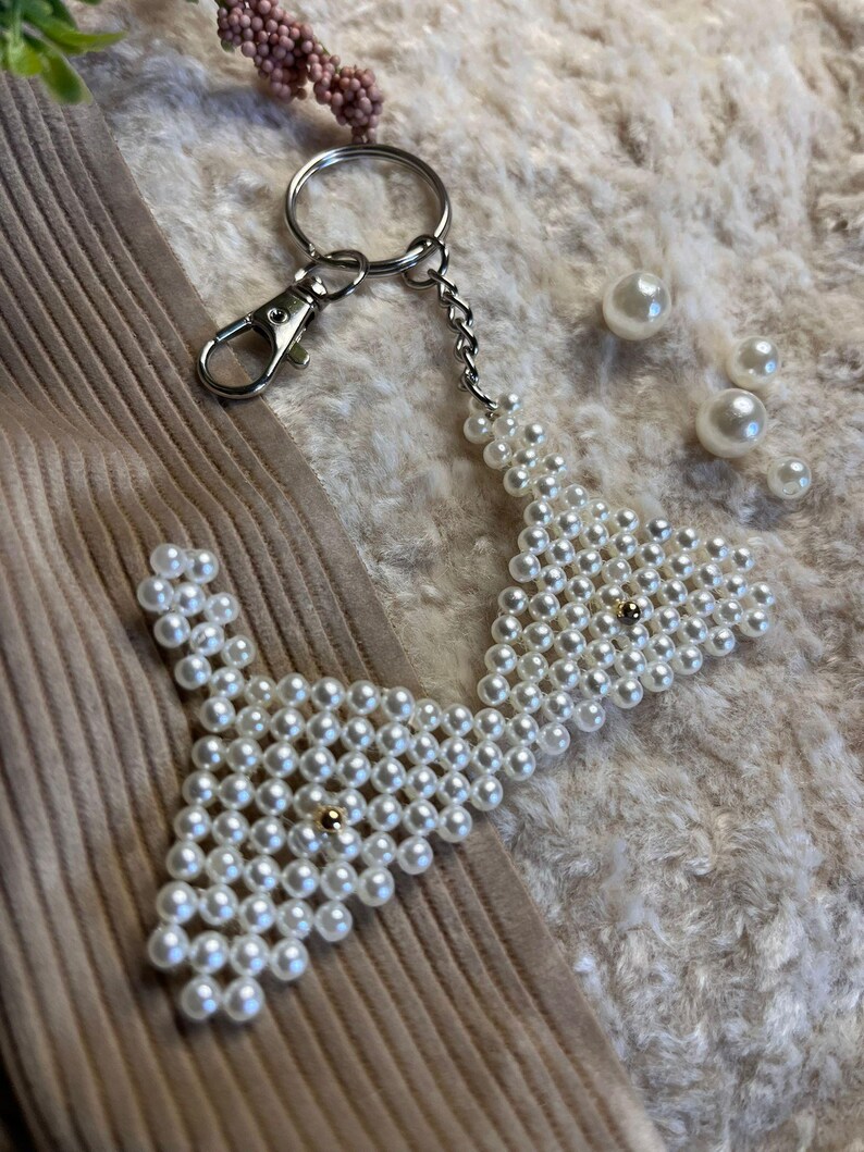 Puede incluir: Un llavero blanco con cuentas en forma de pajarita con un cierre y cadena plateados. El llavero est&aacute; hecho de peque&ntilde;as cuentas blancas redondas y presenta una peque&ntilde;a cuenta dorada en el centro de cada secci&oacute;n de pajarita.