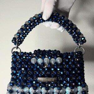 May include: A beaded handbag in shades of blue and white, held by a gloved hand. The bag has a rectangular shape with a handle and silver hardware. The word "CHEMEA" is displayed on a silver plate on the front of the bag.