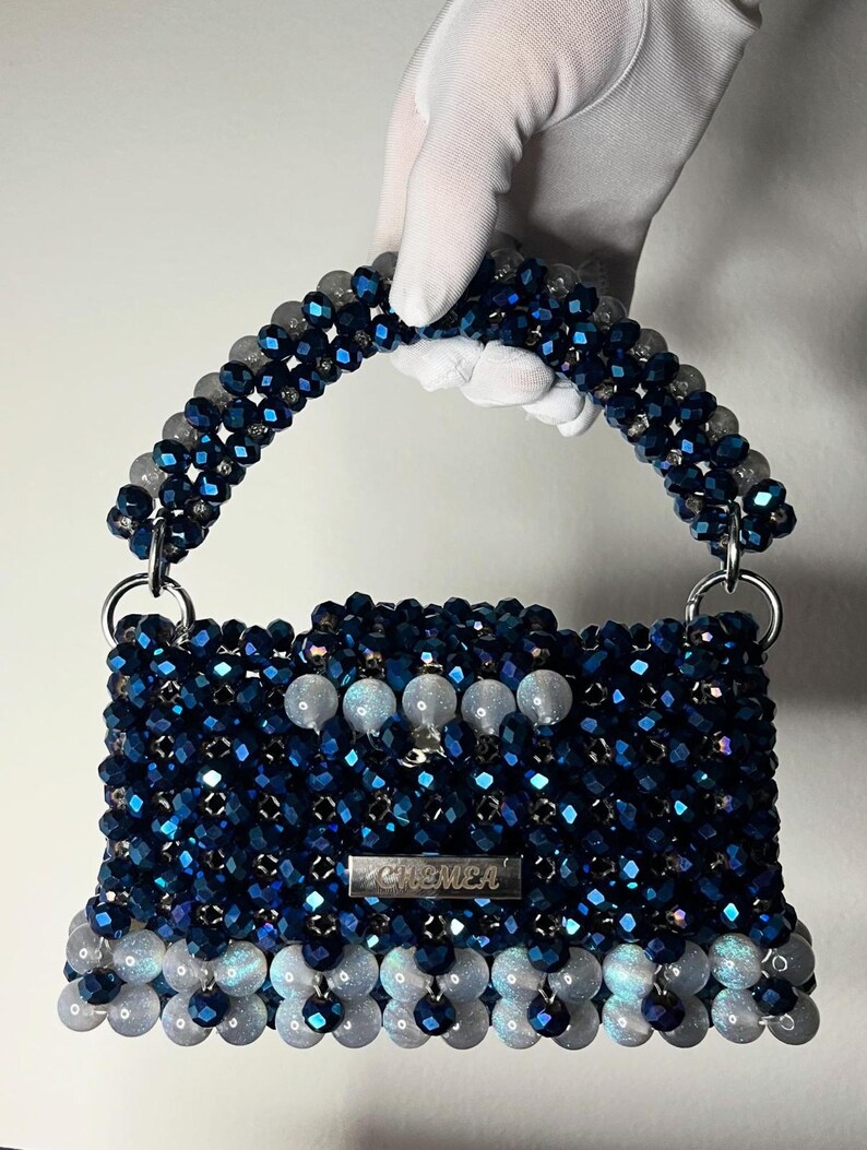 May include: A beaded handbag in shades of blue and white, held by a gloved hand. The bag has a rectangular shape with a handle and silver hardware. The word "CHEMEA" is displayed on a silver plate on the front of the bag.