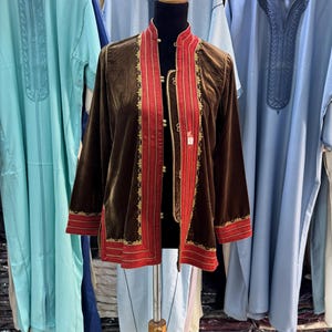 May include: A brown velvet jacket with red and gold detailing, displayed on a mannequin. The jacket features a mandarin collar and long sleeves. Other garments in shades of blue and turquoise are visible.