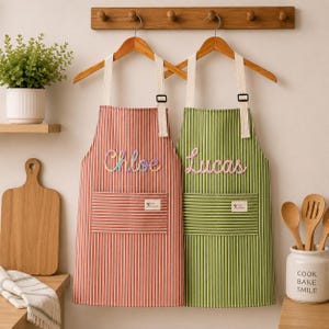 Puede incluir: Dos delantales personalizados colgados de perchas de madera. Uno es rojo con rayas blancas y el nombre "Chloe" en escritura colorida. El otro es verde con rayas blancas y el nombre "Lucas". Ambos tienen un bolsillo y un pequeño logotipo.