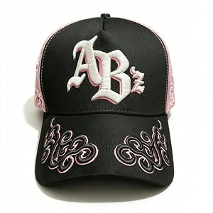 Peut inclure: Casquette de baseball noire avec des d&eacute;tails roses et des broderies. La casquette pr&eacute;sente les lettres "ABz" en blanc, avec des broderies roses sur la visi&egrave;re et les c&ocirc;t&eacute;s. Le design comprend un motif cachemire sur les c&ocirc;t&eacute;s et des volutes d&eacute;coratives sur la visi&egrave;re.