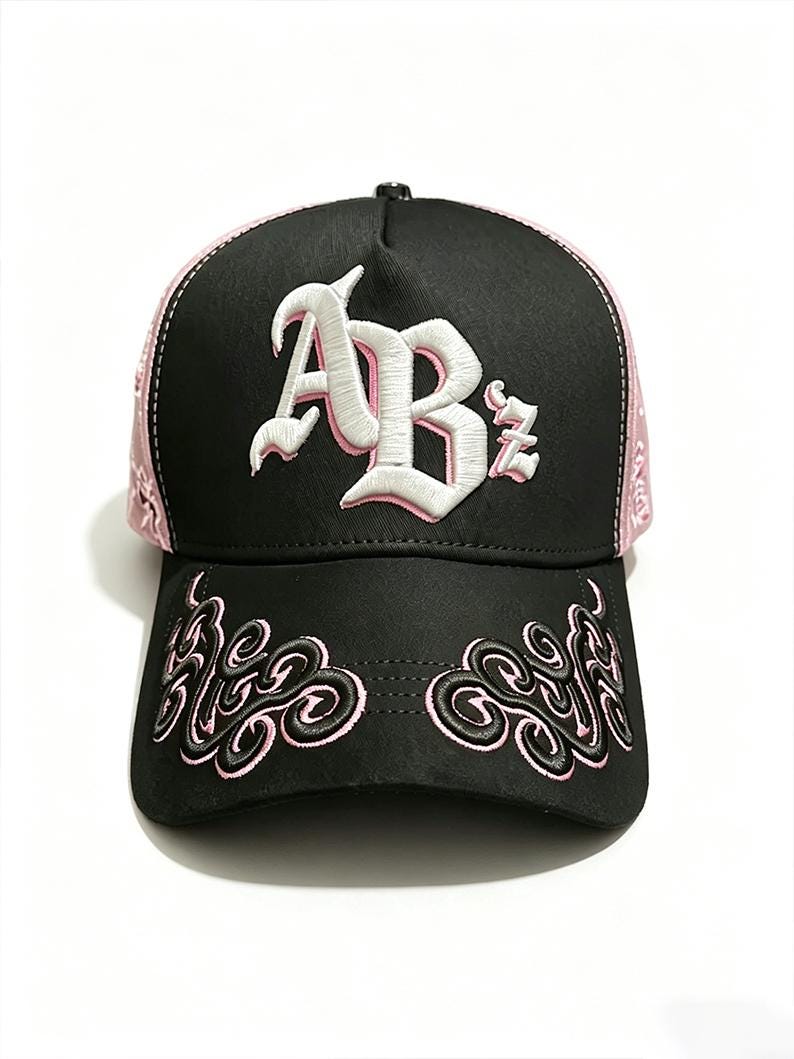 Peut inclure: Casquette de baseball noire avec des d&eacute;tails roses et des broderies. La casquette pr&eacute;sente les lettres "ABz" en blanc, avec des broderies roses sur la visi&egrave;re et les c&ocirc;t&eacute;s. Le design comprend un motif cachemire sur les c&ocirc;t&eacute;s et des volutes d&eacute;coratives sur la visi&egrave;re.