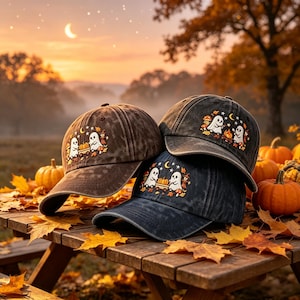 Puede incluir: Tres gorras de béisbol con aspecto desgastado en tonos carbón y marrón oscuro, cada una con un bordado en el panel frontal que muestra dos fantasmas blancos, una calabaza, hojas de otoño, setas, una luna creciente y estrellas.