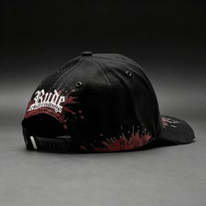 May include: Black baseball cap with a flat brim, featuring the word "Rude" in white script with red accents. The cap has a splatter design in red and a snapback closure.