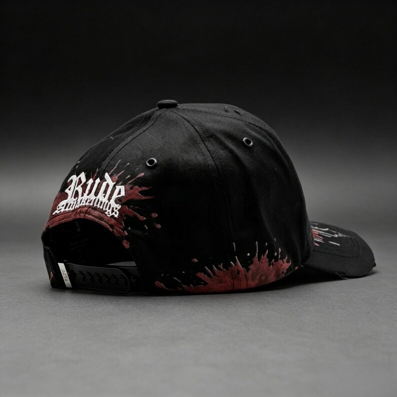 May include: Black baseball cap with a flat brim, featuring the word "Rude" in white script with red accents. The cap has a splatter design in red and a snapback closure.