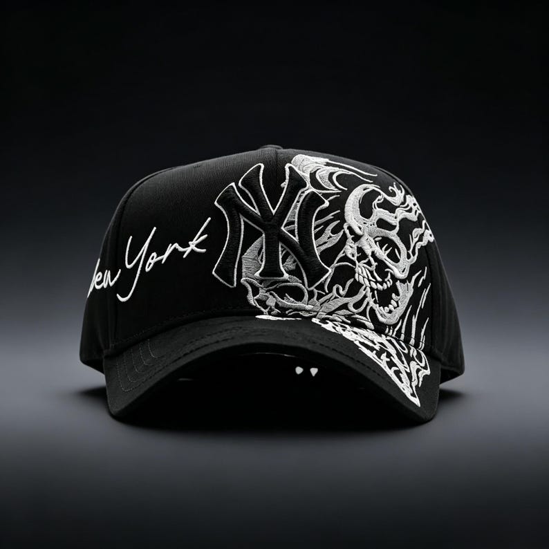 May include: Black baseball cap featuring white embroidered designs. The cap displays the New York Yankees logo and the words "New York" in script. A detailed skull graphic is on the side, adding a bold, edgy aesthetic to the headwear.