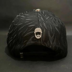 May include: Black baseball cap with a textured leaf pattern. The cap features a silver-colored emblem of a bearded man with a hat and a silver-colored metal clasp. The cap is a stylish accessory.