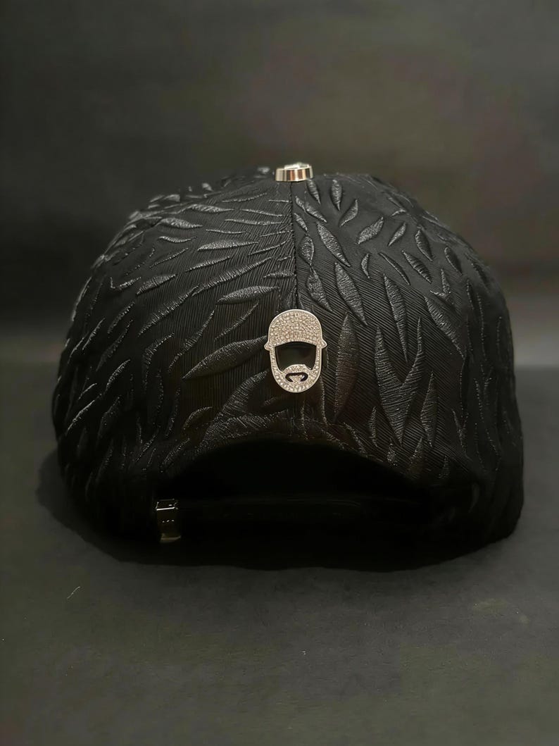 May include: Black baseball cap with a textured leaf pattern. The cap features a silver-colored emblem of a bearded man with a hat and a silver-colored metal clasp. The cap is a stylish accessory.