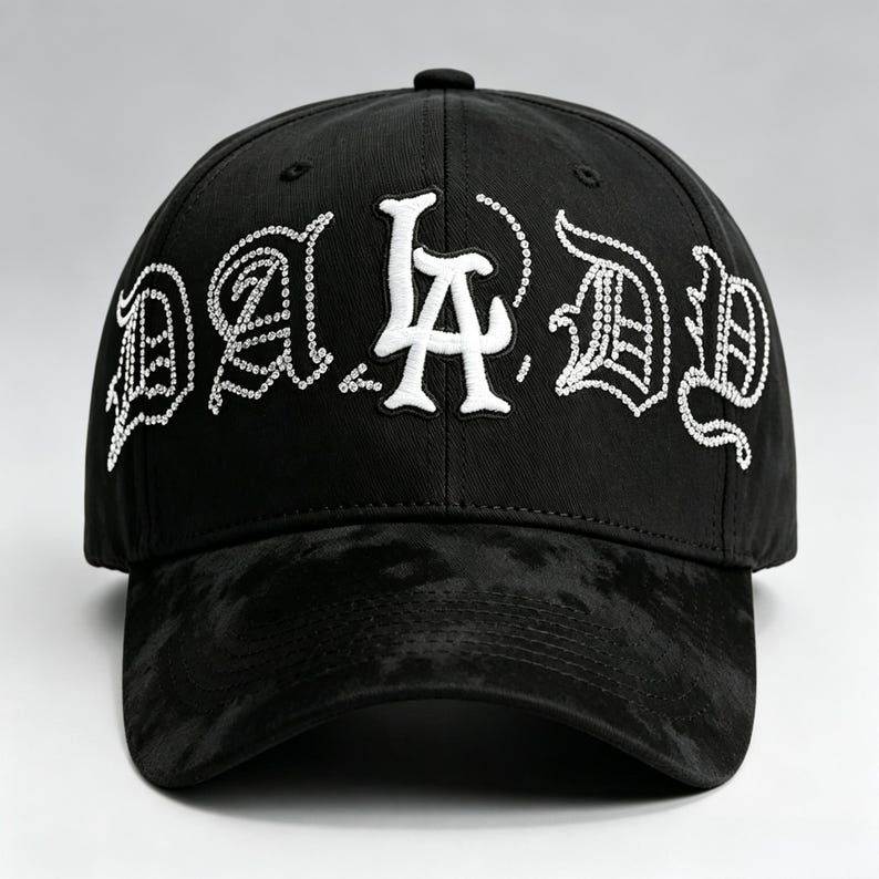 K&ouml;nnte beinhalten: Schwarze Baseballkappe mit dem Text "DA D" in Wei&szlig;, verziert mit Strasssteinen. Der Schirm hat ein dunkles, strukturiertes Muster. Die Kappe ist ein klassisches Sechs-Panel-Design.