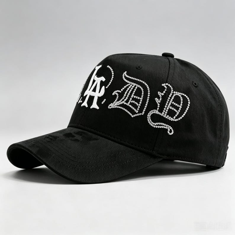 K&ouml;nnte beinhalten: Schwarze Baseballkappe mit wei&szlig;em Strass-Schriftzug "LA DB" in einer stilisierten Schriftart. Die Kappe hat einen gebogenen Schirm und einen Knopf oben. Der Hut ist aus strapazierf&auml;higem Stoff gefertigt.