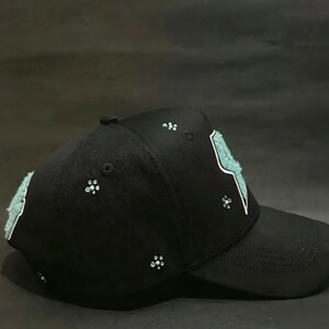May include: Black baseball cap with a curved brim. The cap features a light blue lightning bolt design outlined in white on the front. Small, sparkling embellishments are scattered across the cap, adding a touch of glamour to this accessory.
