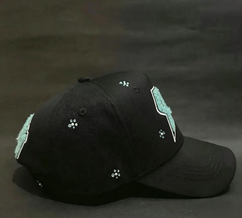 May include: Black baseball cap with a curved brim. The cap features a light blue lightning bolt design outlined in white on the front. Small, sparkling embellishments are scattered across the cap, adding a touch of glamour to this accessory.
