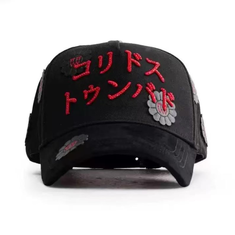 May include: Black baseball cap with red Japanese text and floral patches. The cap has a curved peak and a structured crown. The Japanese text is embroidered in a bold, stylized font. The floral patches are grey and red.