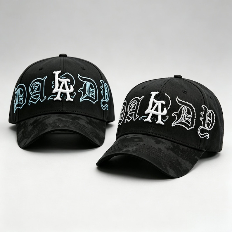 K&ouml;nnte beinhalten: Zwei schwarze Baseballkappen mit dem Wort "DADDY" in Wei&szlig; und Hellblau gestickt. Die Kappen haben ein Camouflagemuster auf Krempe und Krone. Die Kappen werden auf wei&szlig;em Hintergrund pr&auml;sentiert.