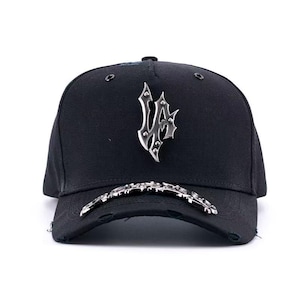 May include: Black baseball cap with a silver metal emblem on the front. The emblem has a gothic design. The brim of the cap has a silver metal design and distressed edges.