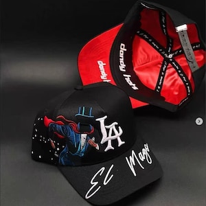 May include: Black baseball cap with a red satin lining and the text "dandy hats". The cap features an embroidered design of a magician with the text "LA" and "El Mago".