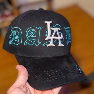 K&ouml;nnte beinhalten: Schwarze Baseballkappe mit "LA" in Wei&szlig; und "DA" in t&uuml;rkisfarbener Stickerei. Die Kappe hat einen gebogenen Schirm und eine strukturierte Krone. Der Hut ist aus einem weichen Material gefertigt.