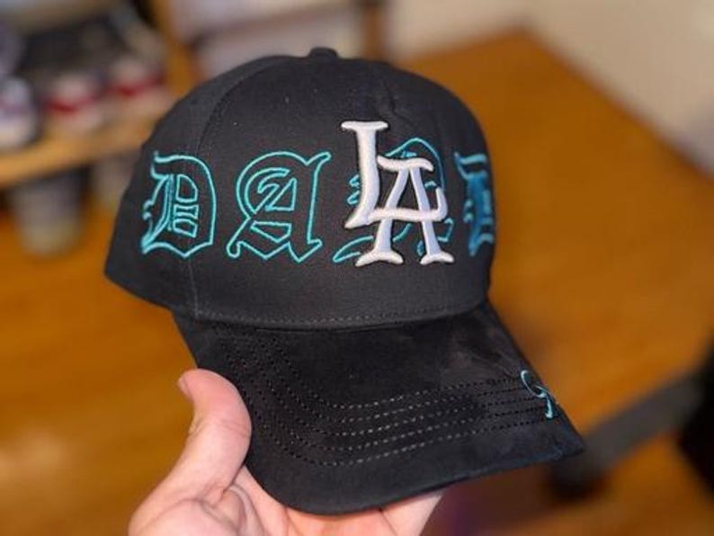 K&ouml;nnte beinhalten: Schwarze Baseballkappe mit "LA" in Wei&szlig; und "DA" in t&uuml;rkisfarbener Stickerei. Die Kappe hat einen gebogenen Schirm und eine strukturierte Krone. Der Hut ist aus einem weichen Material gefertigt.