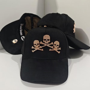 May include: Black baseball caps featuring gold skull and crossbones designs. One cap has the word "Darbas" embroidered on the side. The caps have a curved peak and a structured design.