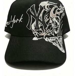 May include: Black baseball cap with white embroidered design. The cap features the text "New York" in cursive and a stylized skull and "NY" logo. The cap has a curved brim and a button on top.