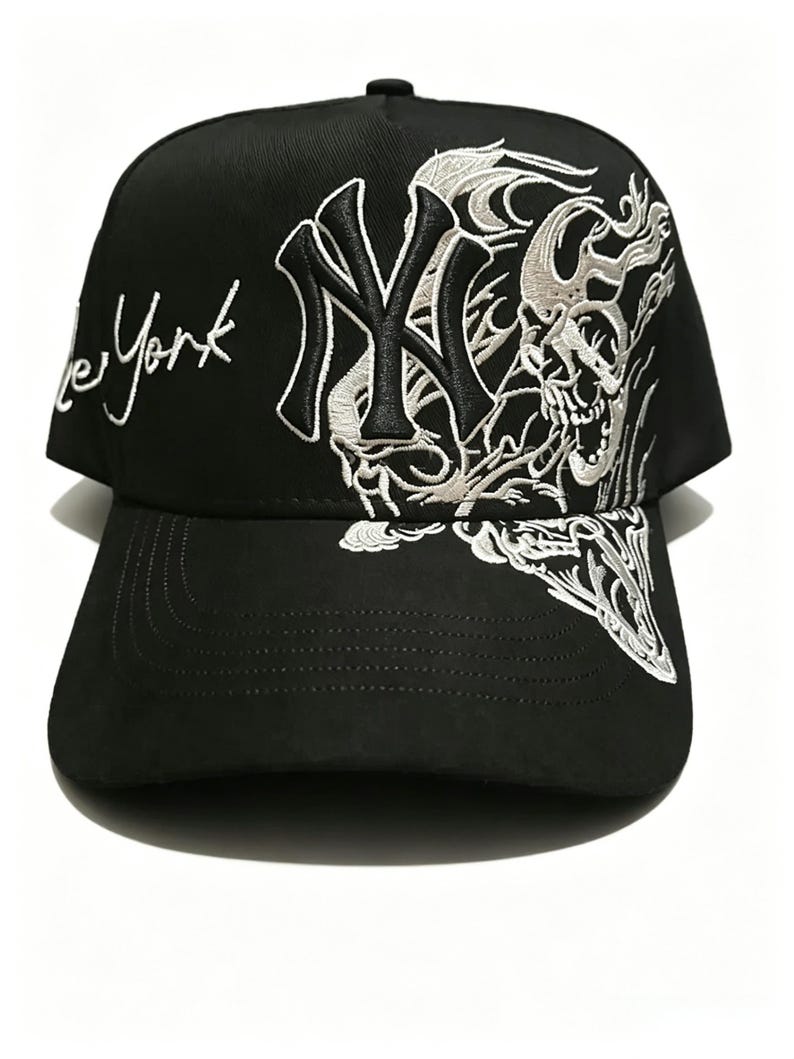 May include: Black baseball cap with white embroidered design. The cap features the text "New York" in cursive and a stylized skull and "NY" logo. The cap has a curved brim and a button on top.