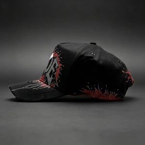 May include: Black baseball cap with a distressed brim, featuring a white and gray graphic design with red splatter accents. The cap has a curved bill and a structured crown, suitable for casual wear.