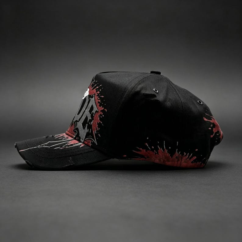 May include: Black baseball cap with a distressed brim, featuring a white and gray graphic design with red splatter accents. The cap has a curved bill and a structured crown, suitable for casual wear.