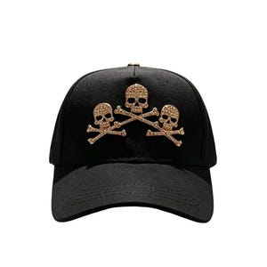 May include: Black baseball cap featuring three gold-colored skull and crossbones designs. The skulls are embellished with small, sparkling details. The cap has a curved brim and a structured crown, suitable for casual wear.