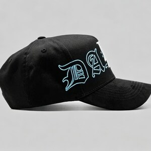 K&ouml;nnte beinhalten: Schwarze Baseballkappe mit dem Wort "DA" in einem stilisierten Schriftzug, der hellblau umrandet ist. Die Kappe hat einen gebogenen Schirm und einen Knopf oben. Der Hintergrund ist einfarbig.
