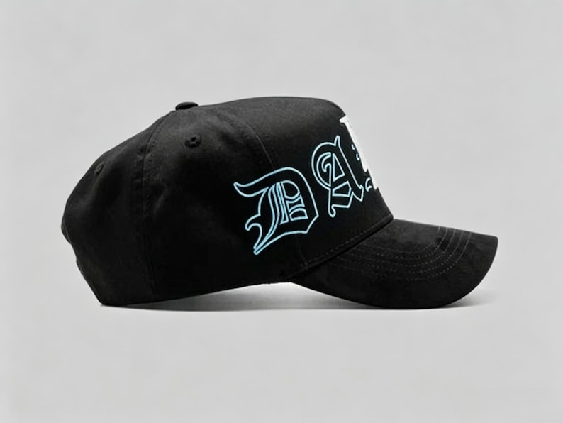 K&ouml;nnte beinhalten: Schwarze Baseballkappe mit dem Wort "DA" in einem stilisierten Schriftzug, der hellblau umrandet ist. Die Kappe hat einen gebogenen Schirm und einen Knopf oben. Der Hintergrund ist einfarbig.